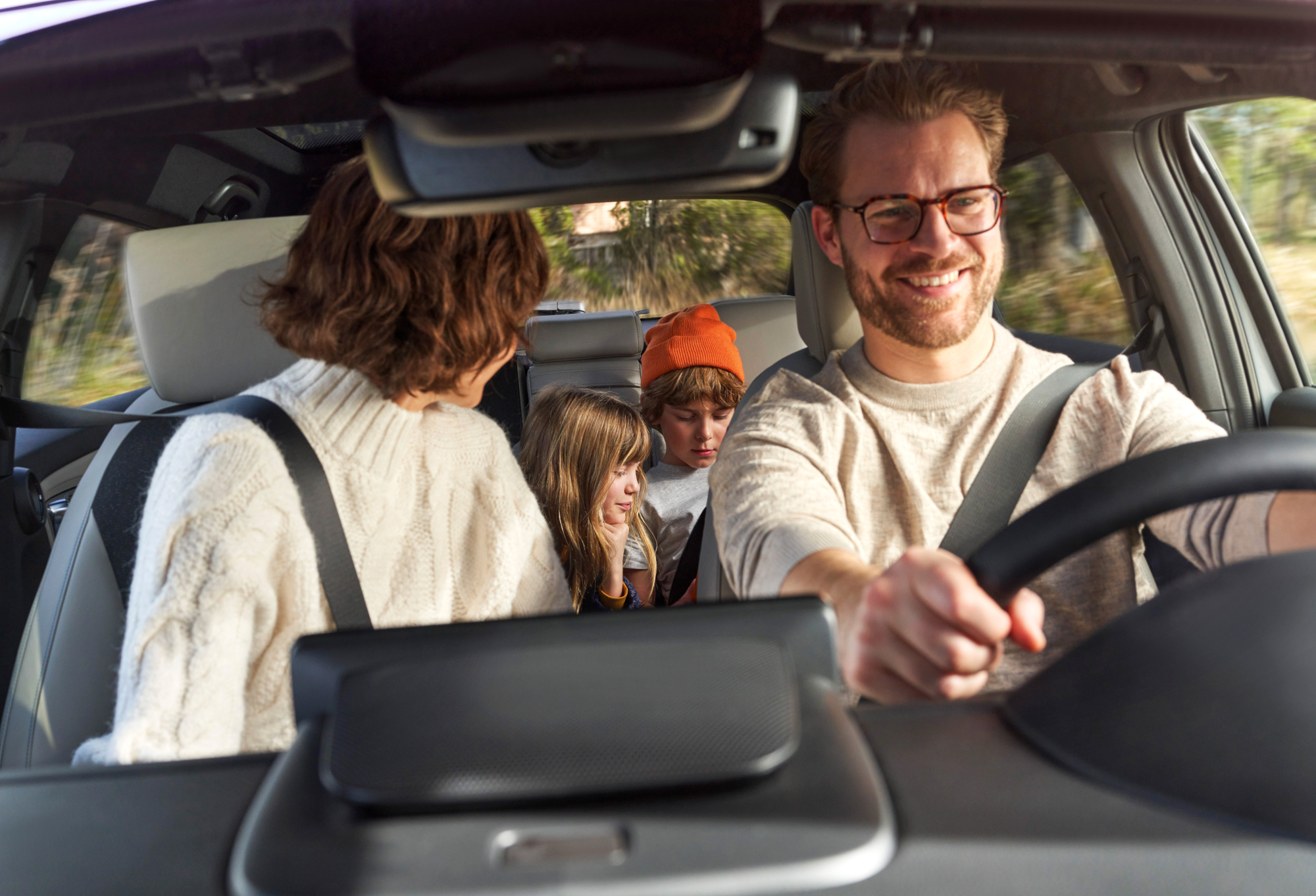 Interior view of a Honda vehicle with a family on a drive. The driver, wearing glasses and a light-colored sweater, smiles while steering. In the backseat, three children are visible, one wearing an orange hat. The car's spacious interior and dashboard are partially shown.